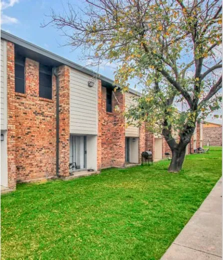Corner of a brick and siding apartment building beside a tree, a green lawn, and a concrete walkway.