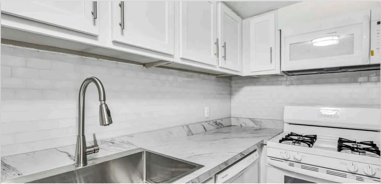 Apartment kitchen with white cabinets, a white subway-tile backsplash, a marble-look countertop, a stainless steel sink with a pull-down faucet, and a white gas range with a microwave above it.