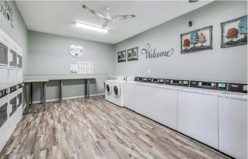 On-site clothes care center, a bright room with a row of washers and dryers, wood-look flooring, a ceiling fan, and a Welcome sign with framed art on the wall.